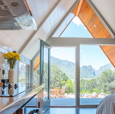 Bedsit with a peaked roof framed glass wall with an open door, a view of wooden patio furniture, mountains and greenery backdrop